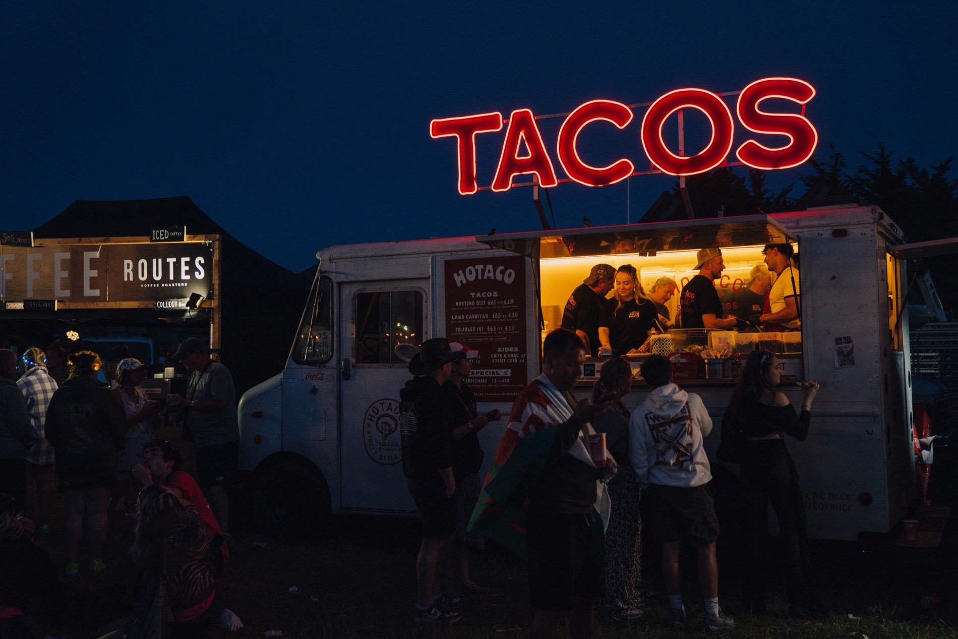 The step van at night, the neon TACOS sign lit red above it, the crew working the lit hatch and a crowd in front.