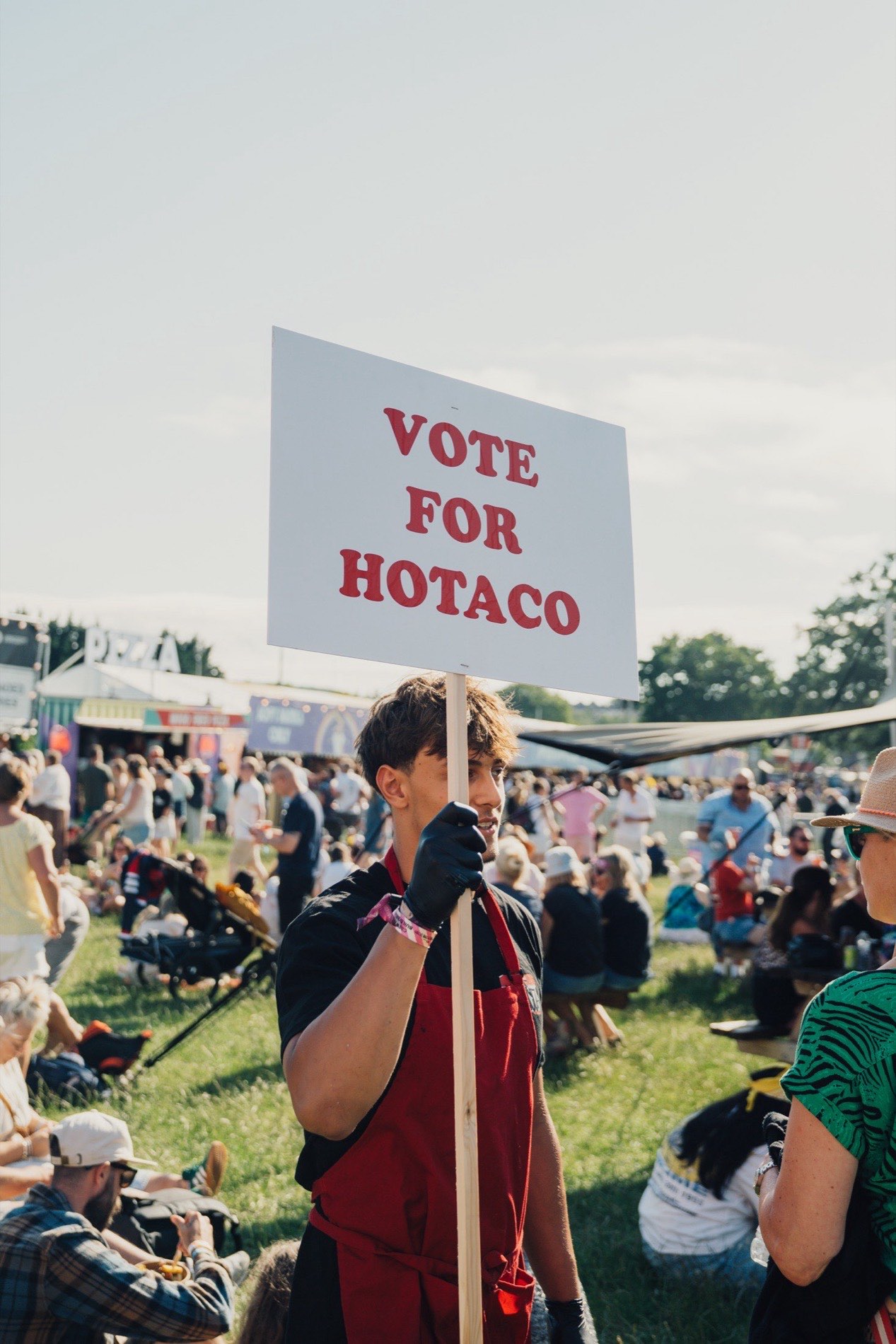A member of the HoTaco crew holding a Vote For HoTaco placard in a busy festival crowd.