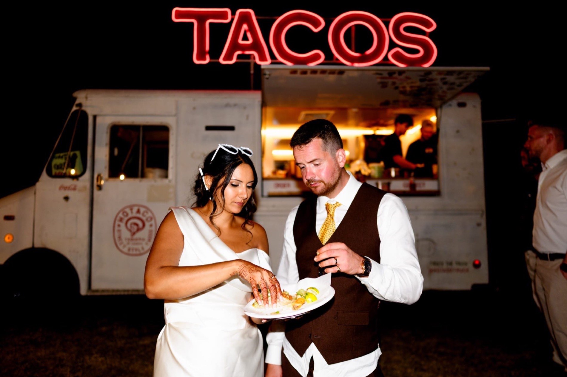 A bride and groom stood together at the HoTaco truck at night, eating tacos under the lit TACOS sign.