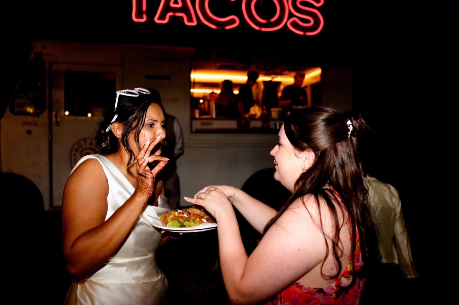 The bride laughing with a guest, both holding plates of tacos in front of the truck.