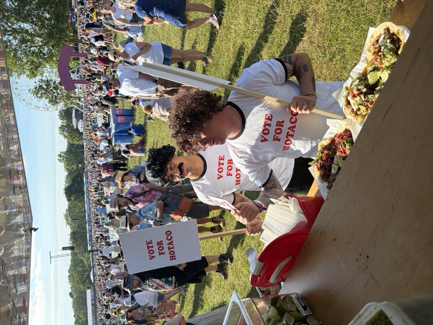 A festival crowd stretching across the field in front of the truck, with a Vote For HoTaco placard held up in the middle of it.
