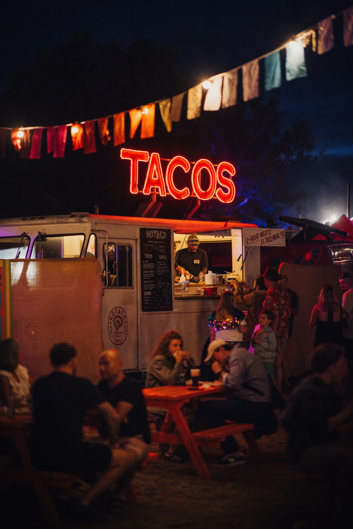 The truck lit up in the evening under the TACOS sign, bunting overhead and a crowd at the tables.