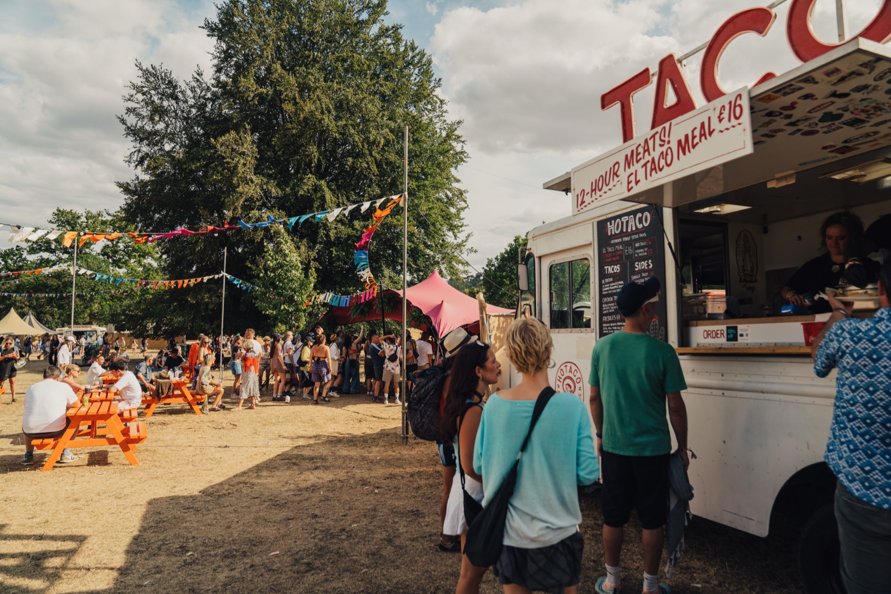 A queue at the truck on a festival site in daylight, the red TACOS sign above the hatch, bunting and picnic tables behind.